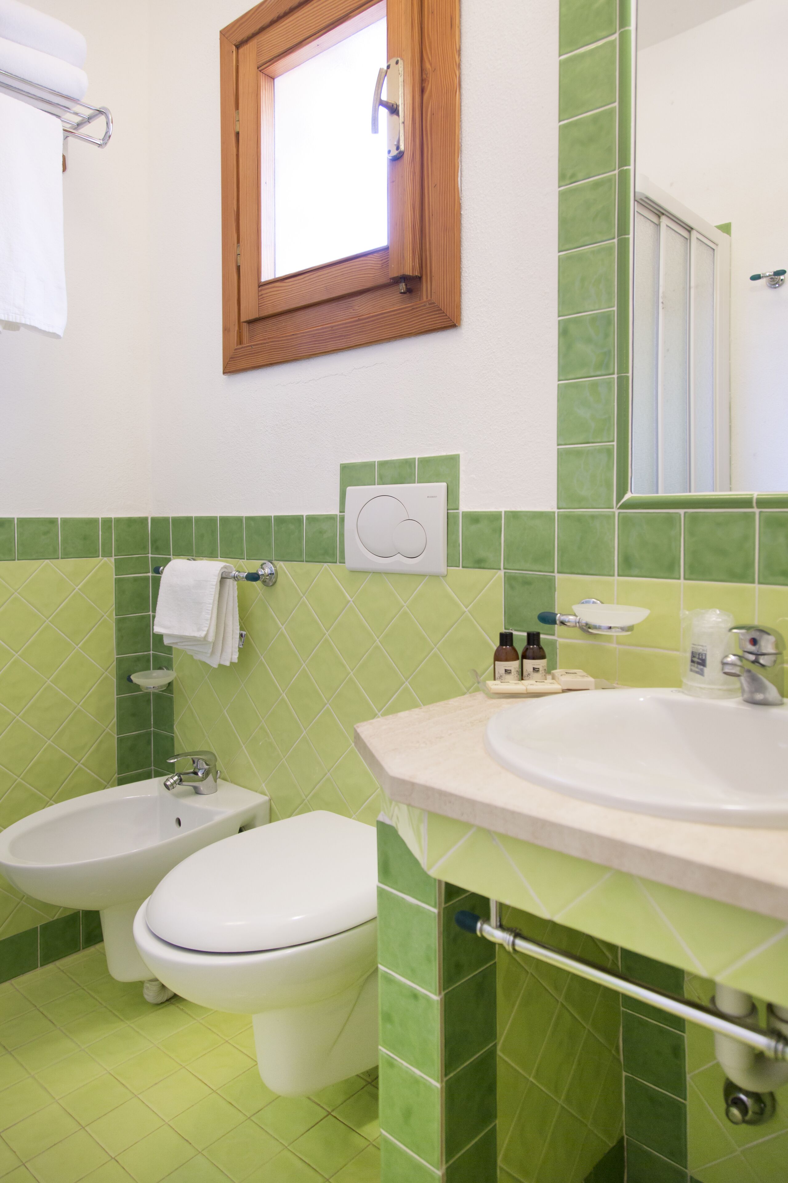 A bathroom with a bidet, toilet, and sink, all in a green-tiled room with a window above the sink.
