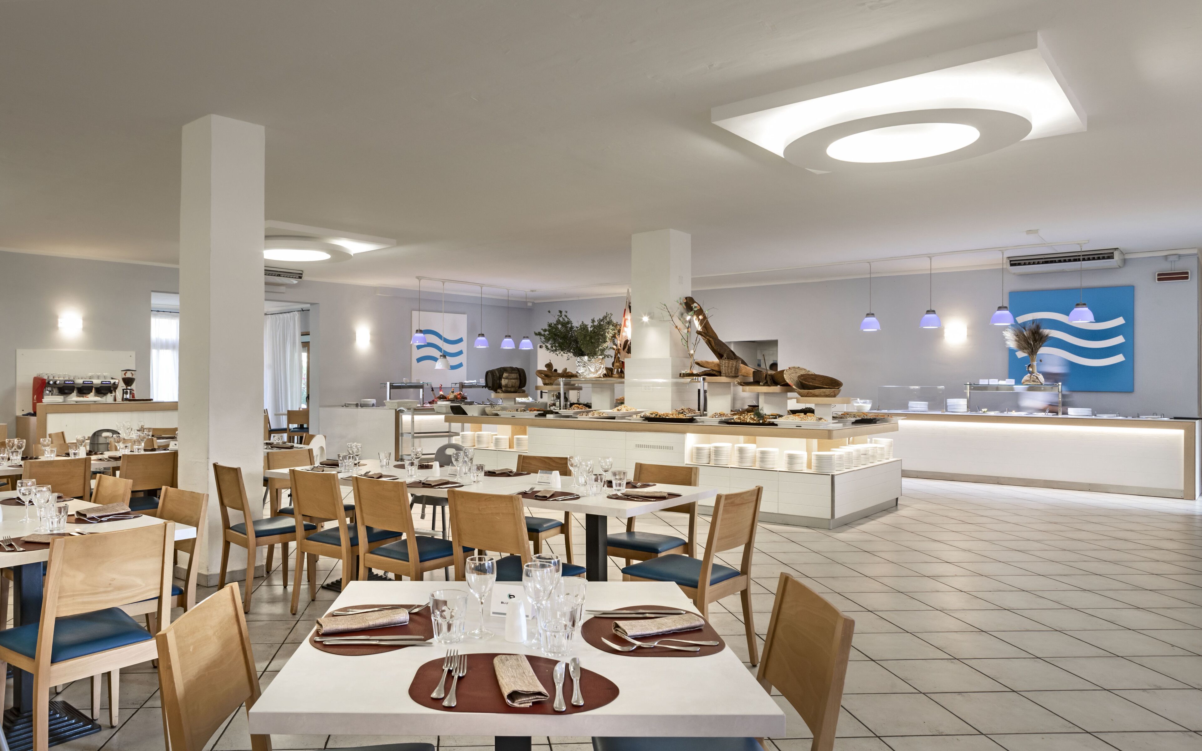 Een modern restaurant met witte tafels, blauwe stoelen met kussens en een centraal gelegen bar-eiland.