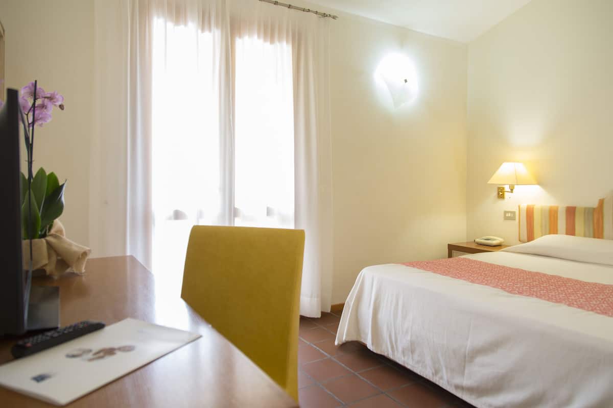 A hotel room with a bed, a desk with a lamp, a chair, and a window with curtains.