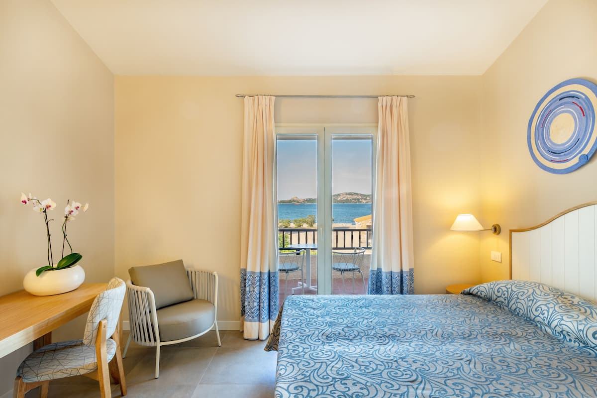 A bedroom with a bed, a chair, a table, and a view of the sea through a glass door.