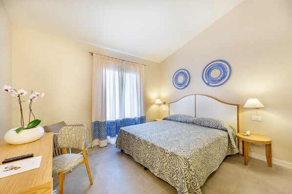 Un dormitorio con cama, escritorio con silla, un florero con flores y ventana con cortinas.