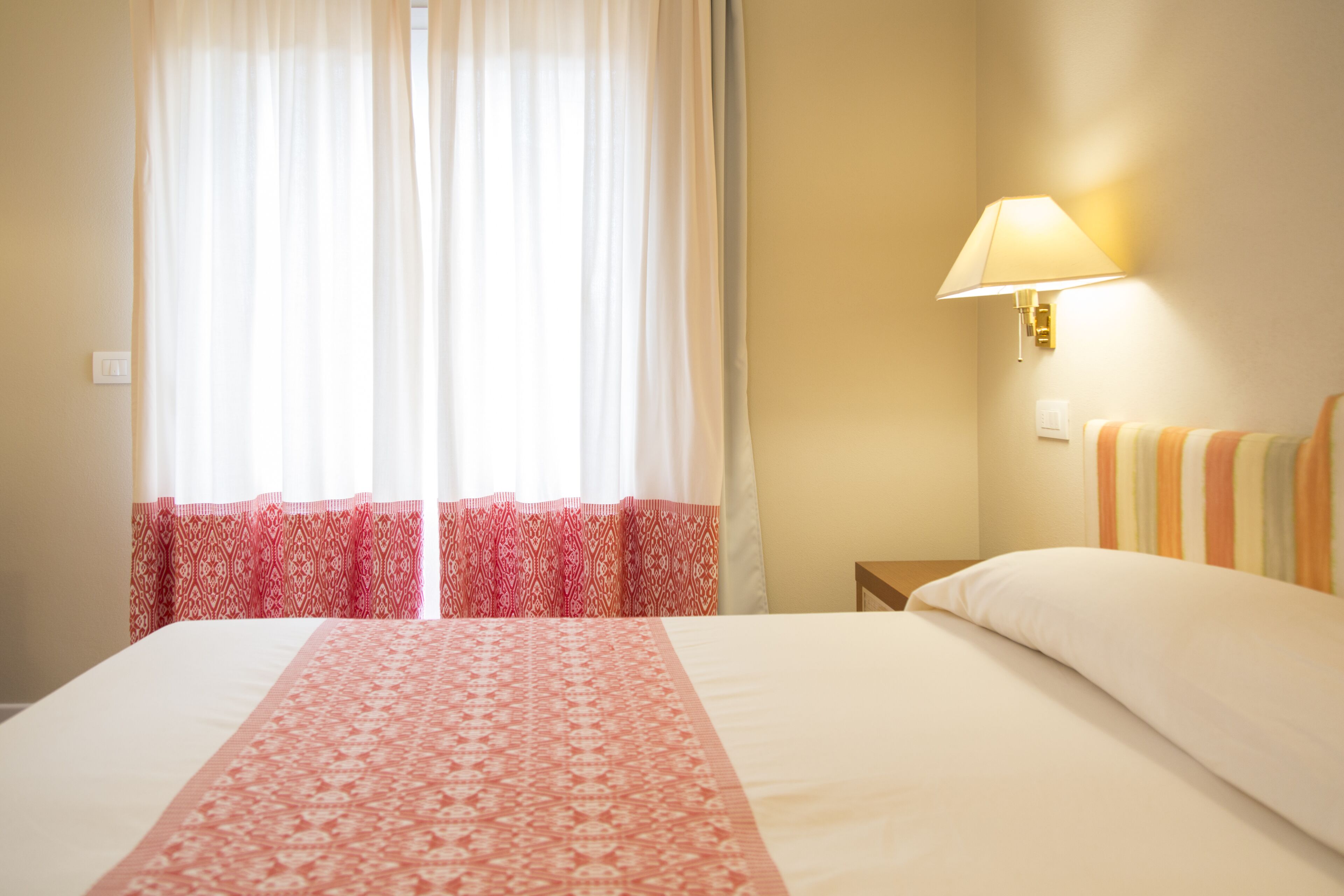 Una habitación de hotel con cama, mesita de noche, lámpara y ventana con cortinas.