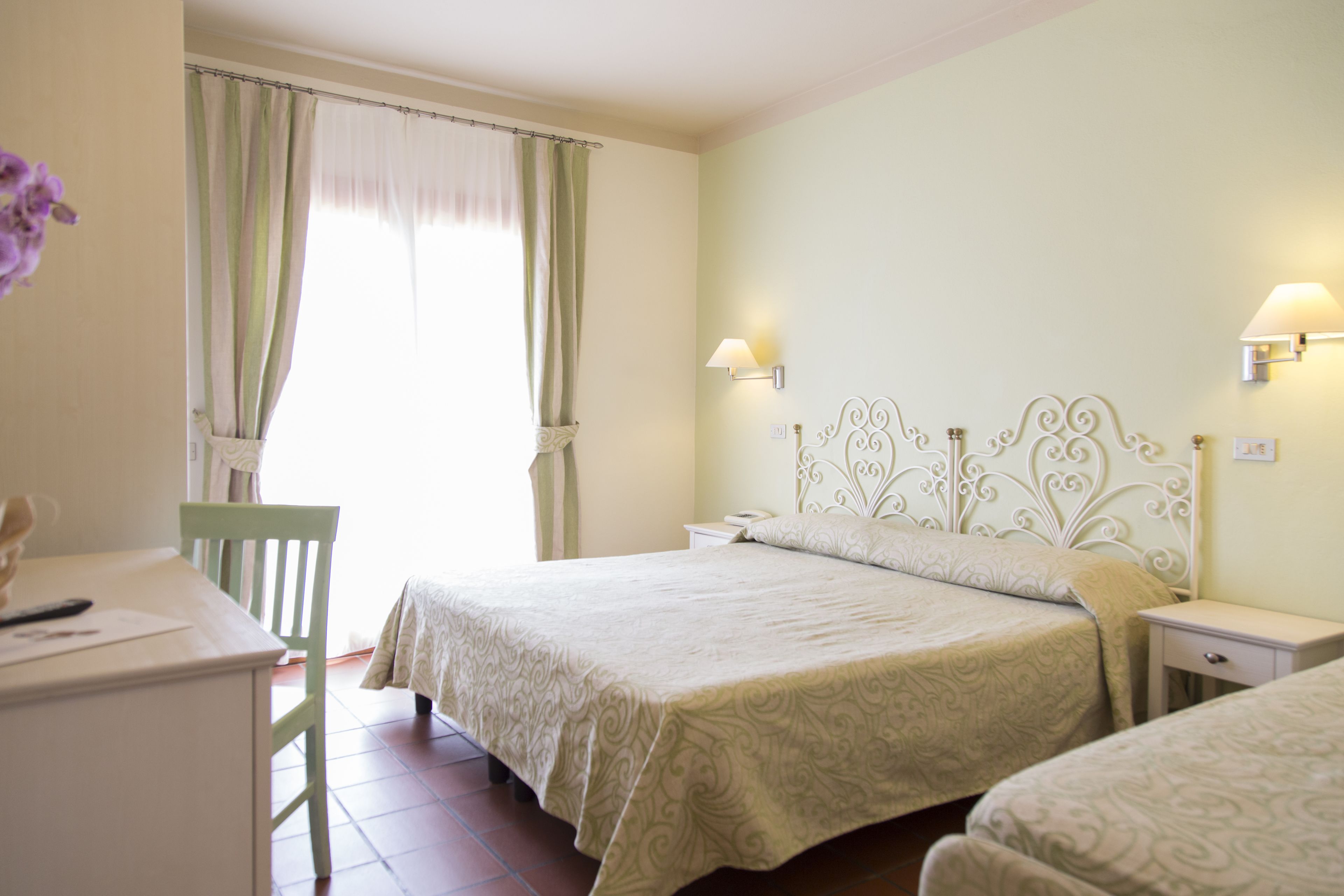 A bedroom with two beds, a desk, and a chair. There are two bedside lamps and a window with curtains.