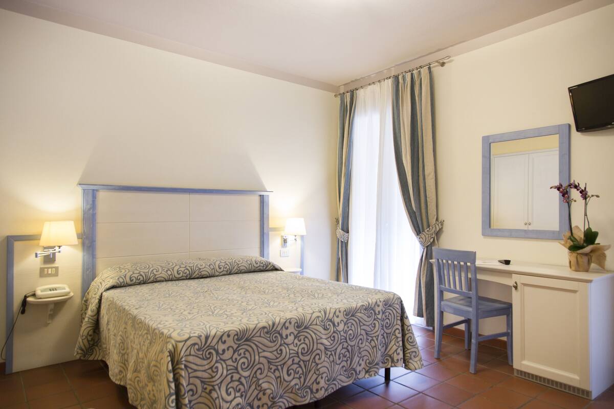 A hotel room with a bed, a desk, a chair, a telephone, and a television on the wall.