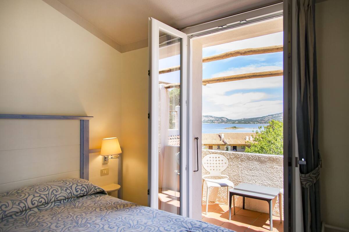 A bedroom with a bed, a nightstand, a lamp, and a sliding glass door leading to a balcony with a chair and a table.