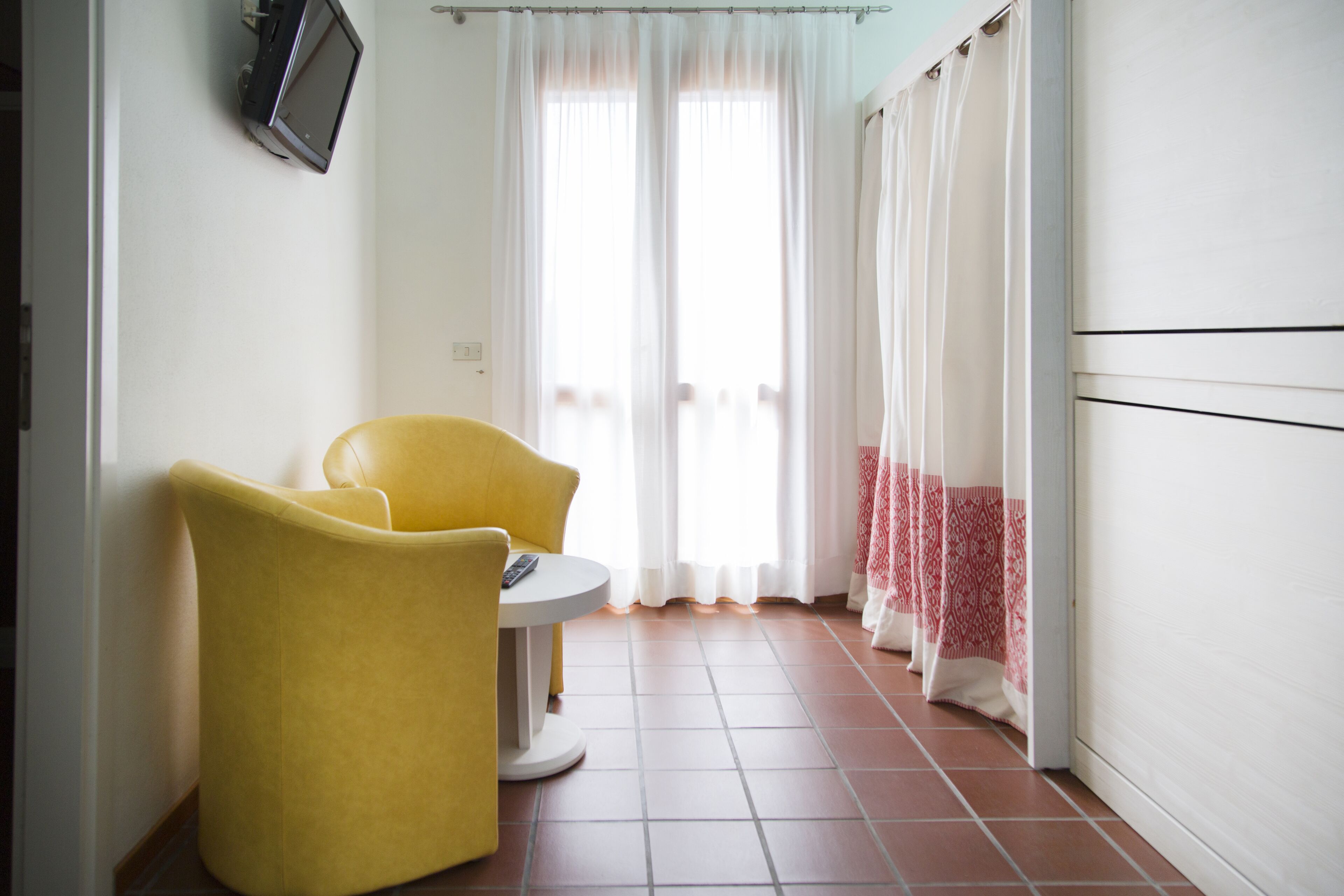 Een kamer met een geel fauteuil, een klein rond tafeltje, een aan de muur bevestigde televisie en een balkon met witte gordijnen.