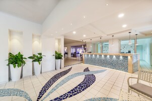 A modern hotel lobby with a reception desk, potted plants, and a tiled floor with a blue and white pattern.