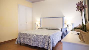 Een slaapkamer met een bed, nachtkastje, lamp en een vaas met bloemen.