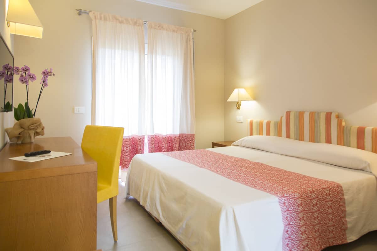 A hotel room with a bed, a desk, a chair, a lamp, and a window with curtains.