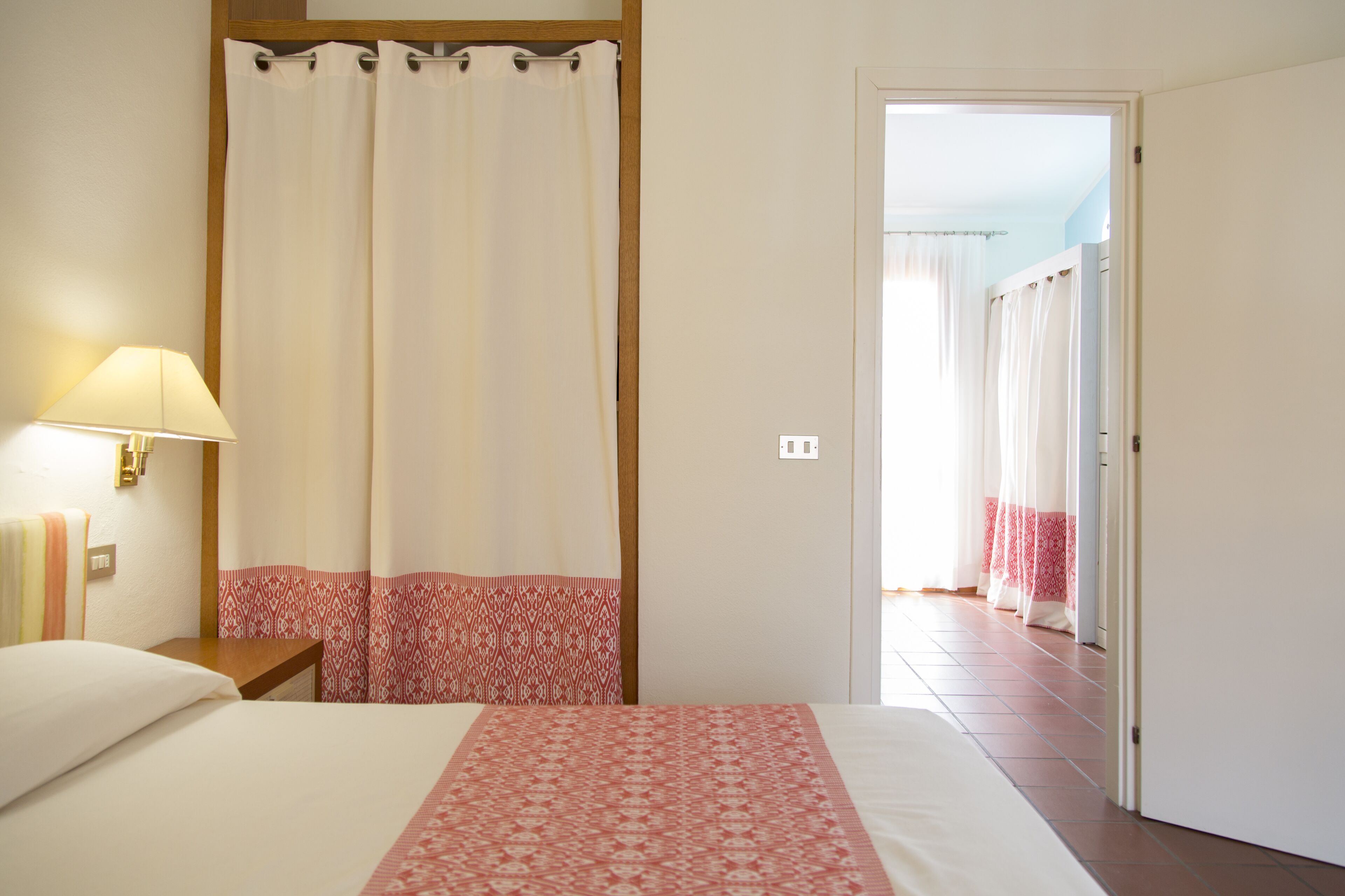 A hotel room with a bed, a bedside table with a lamp, a patterned bedspread, and a view of the corridor with a door leading outside.