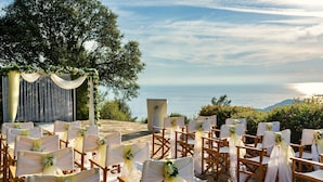 Un lieu de réception en plein air pour les mariages, avec des rangées de chaises en bois et des chaises recouvertes de tissu blanc, le tout mis en valeur par une vue panoramique sur l’océan.