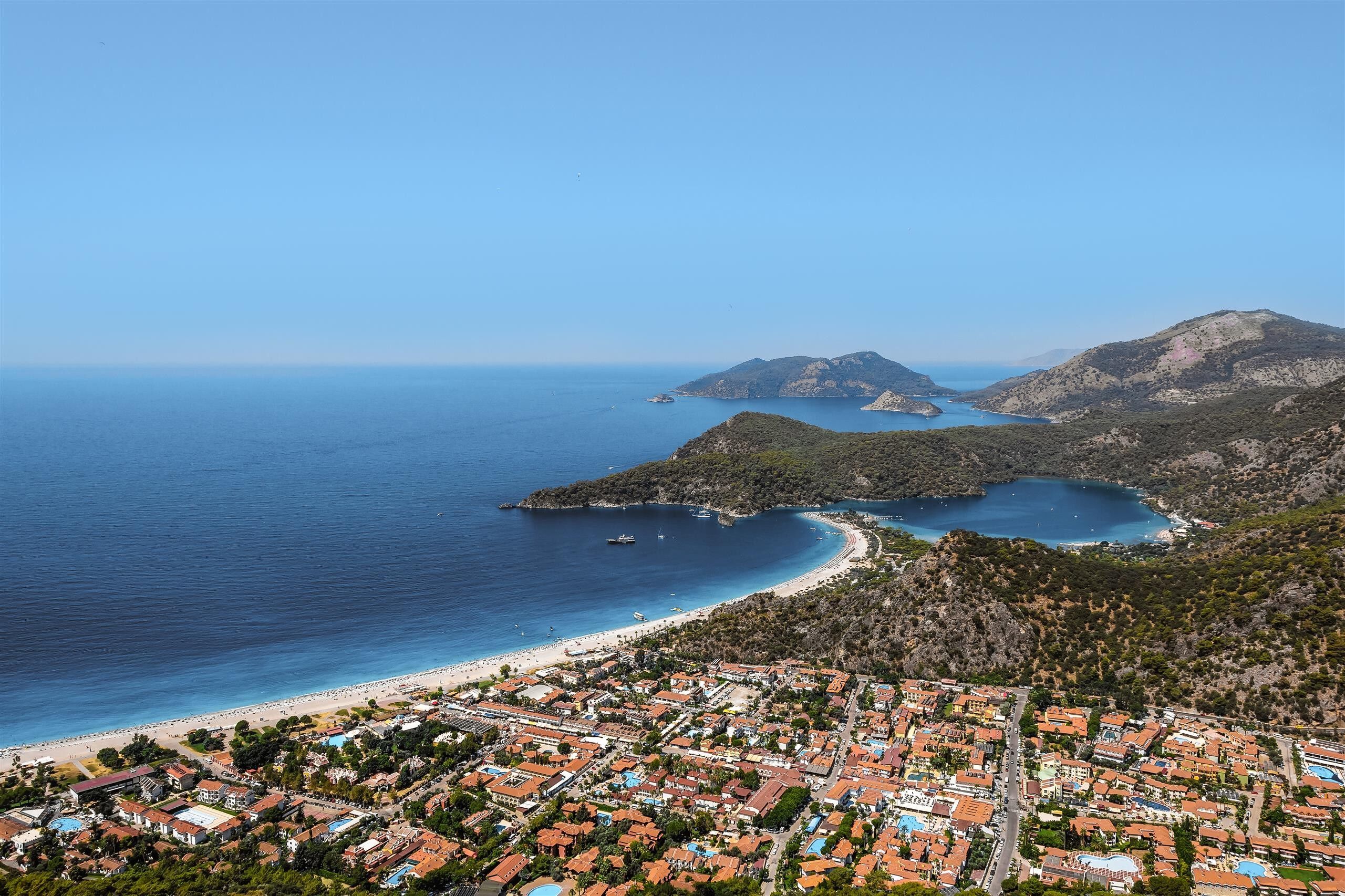 Vista aérea de un pueblo costero con playa, zona residencial y terreno montañoso.