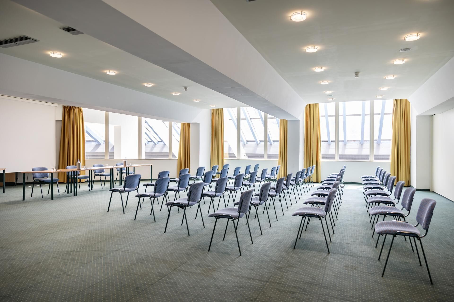 Una sala conferenze con file di sedie, un lungo tavolo e ampie finestre con tende.
