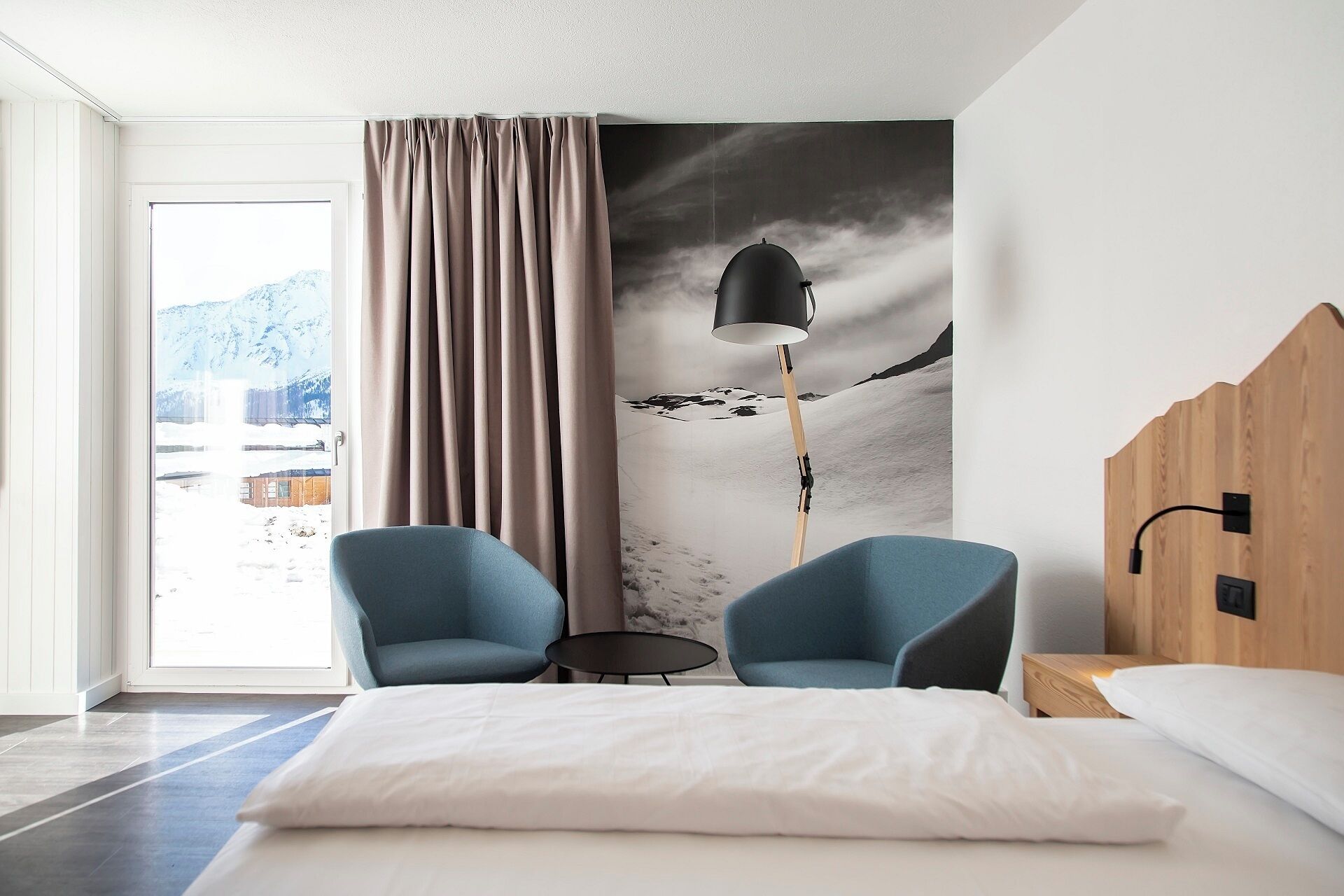 A modern hotel room with a bed, two armchairs, a small table, a lamp, and a view of a snowy landscape through a window.