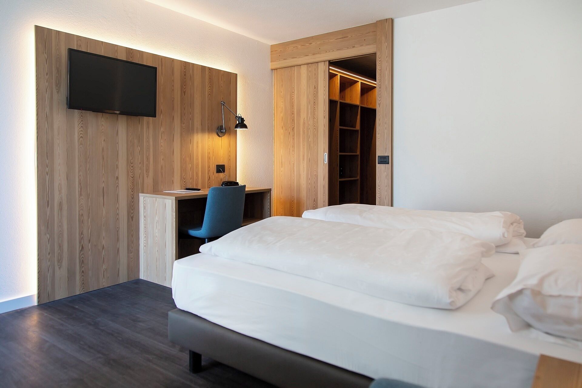 A hotel room with a bed, a desk, a chair, a TV mounted on a wooden wall, and a wardrobe with a mirror.