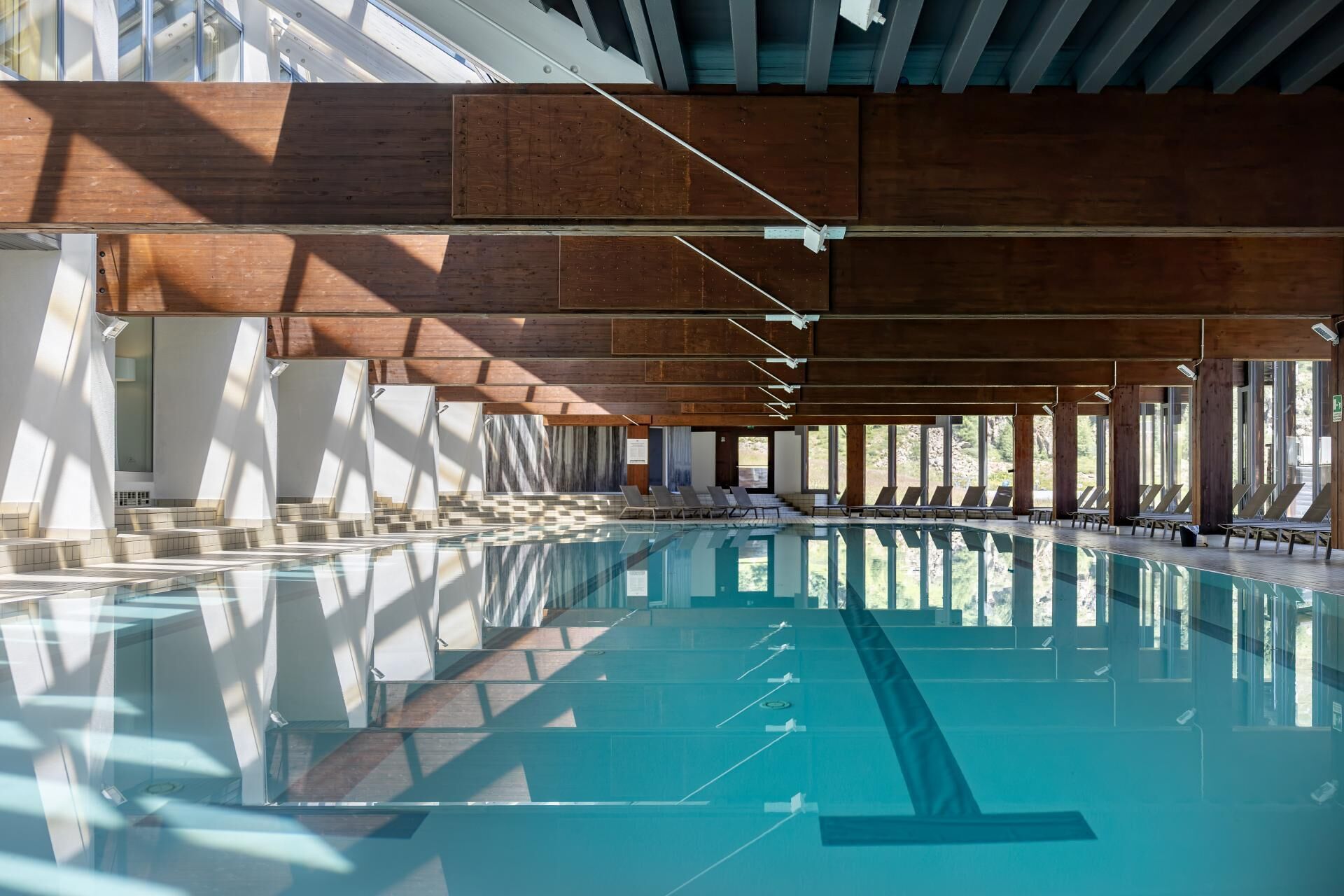 Una amplia piscina interior con agua cristalina, rodeada de espacios iluminados por el sol con vigas de madera y grandes ventanales.