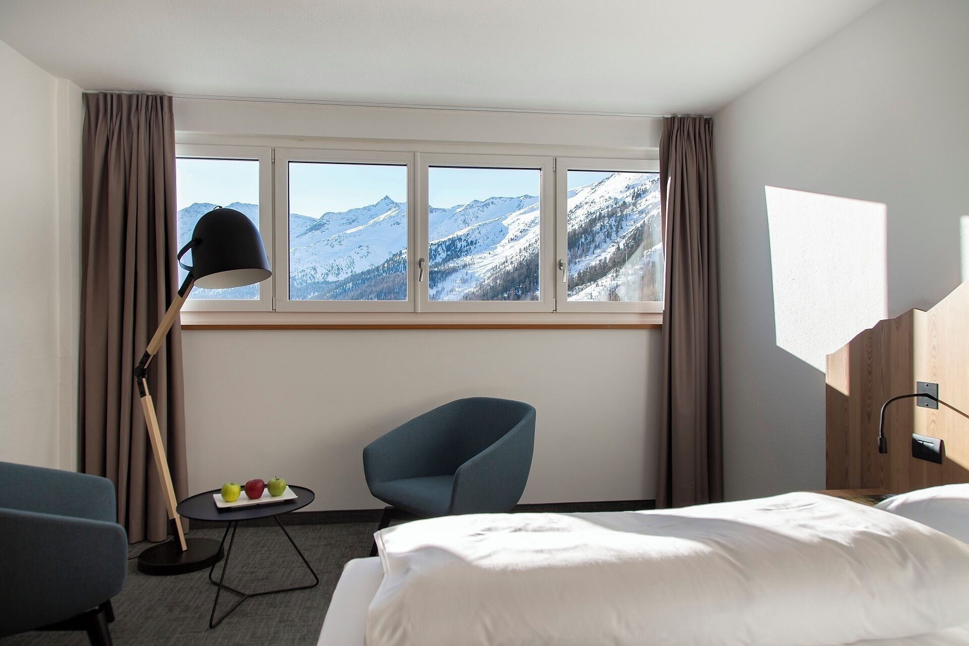 Une chambre d’hôtel avec un lit, une chaise, une petite table avec des pommes, une lampe et une fenêtre donnant sur la montagne.