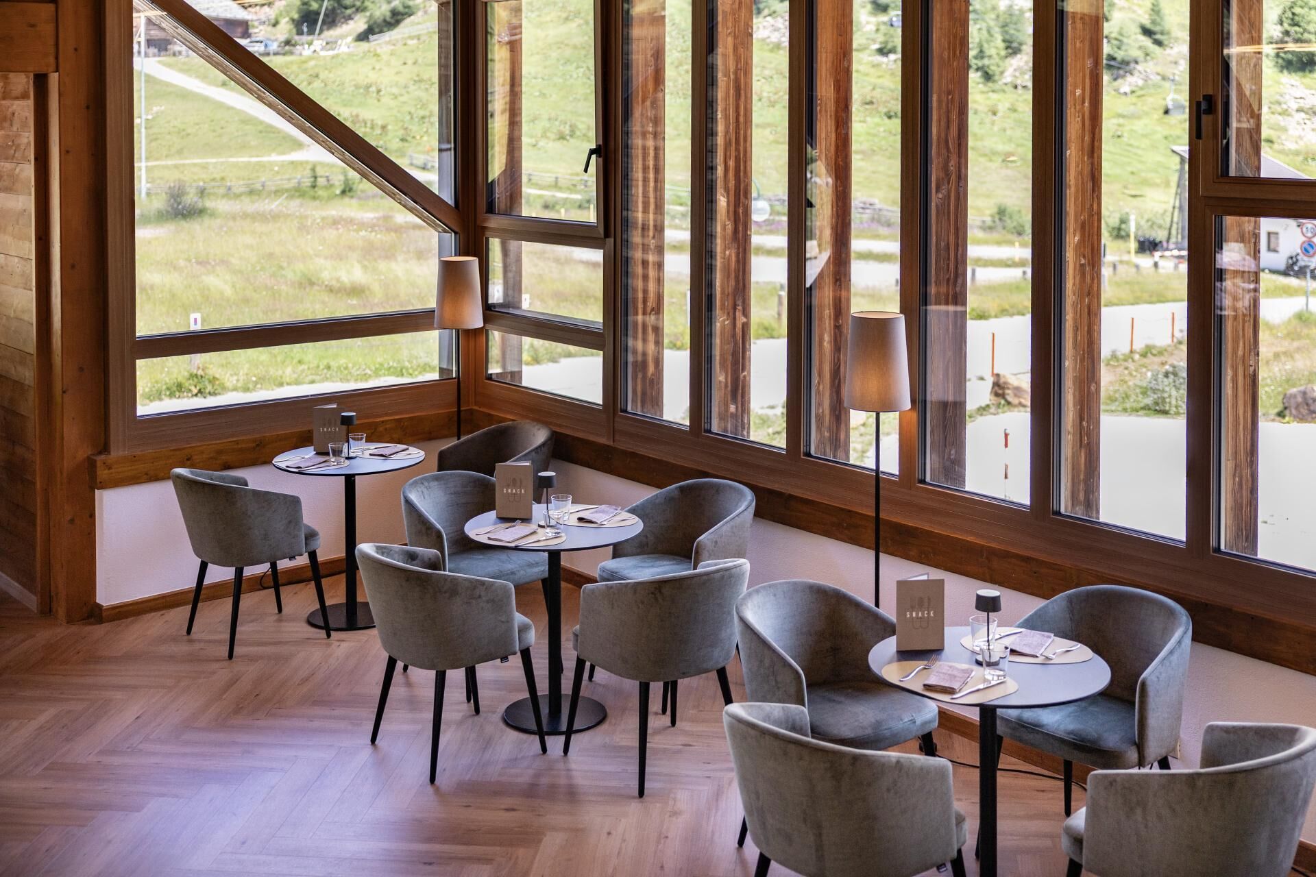 Ein Restaurantinneres mit runden Tischen, grau gepolsterten Stühlen und Blick auf eine grüne Landschaft durch große Fenster.