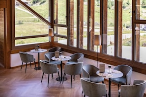 A restaurant interior with round tables, grey upholstered chairs, and a view of a green landscape through large windows. - Blu Senales Active & Family Resort (Senales)