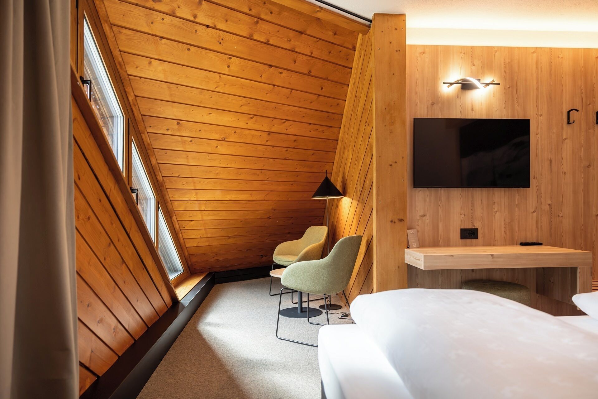 Ein Hotelzimmer mit schräg versiegelter Decke, Holzvertäfelung, einem flachbildfernseher, der über einer Holzkonsole angebracht ist, zwei Sessel und ein Bett.
