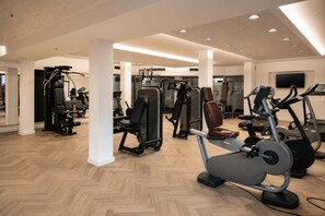 A gym with various exercise machines, a large mirror, and a television screen. - Blu Senales Active & Family Resort (Senales)