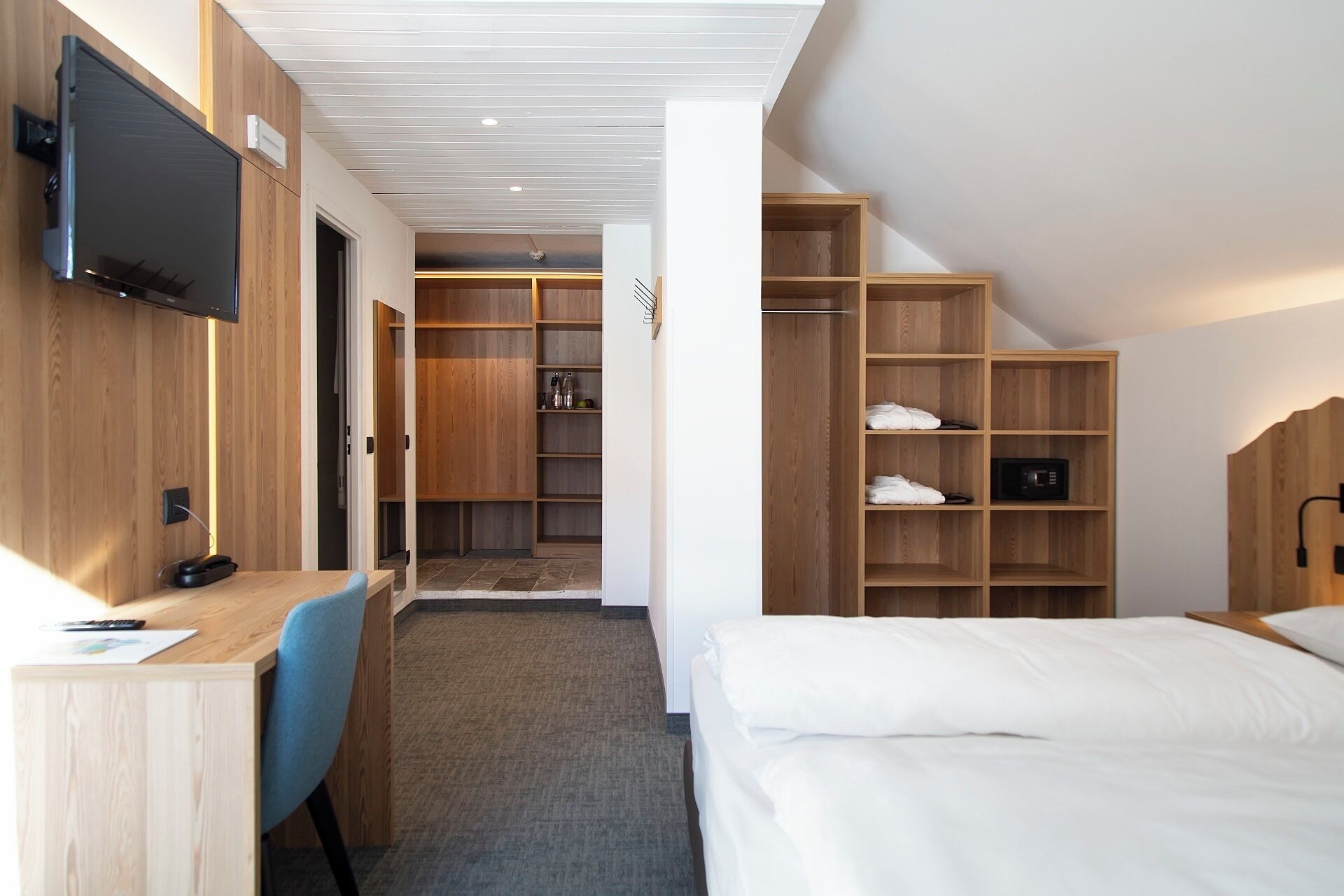Une chambre d’hôtel comprenant un lit, un bureau avec une chaise, un téléviseur fixé au mur et des étagères en bois intégrées.