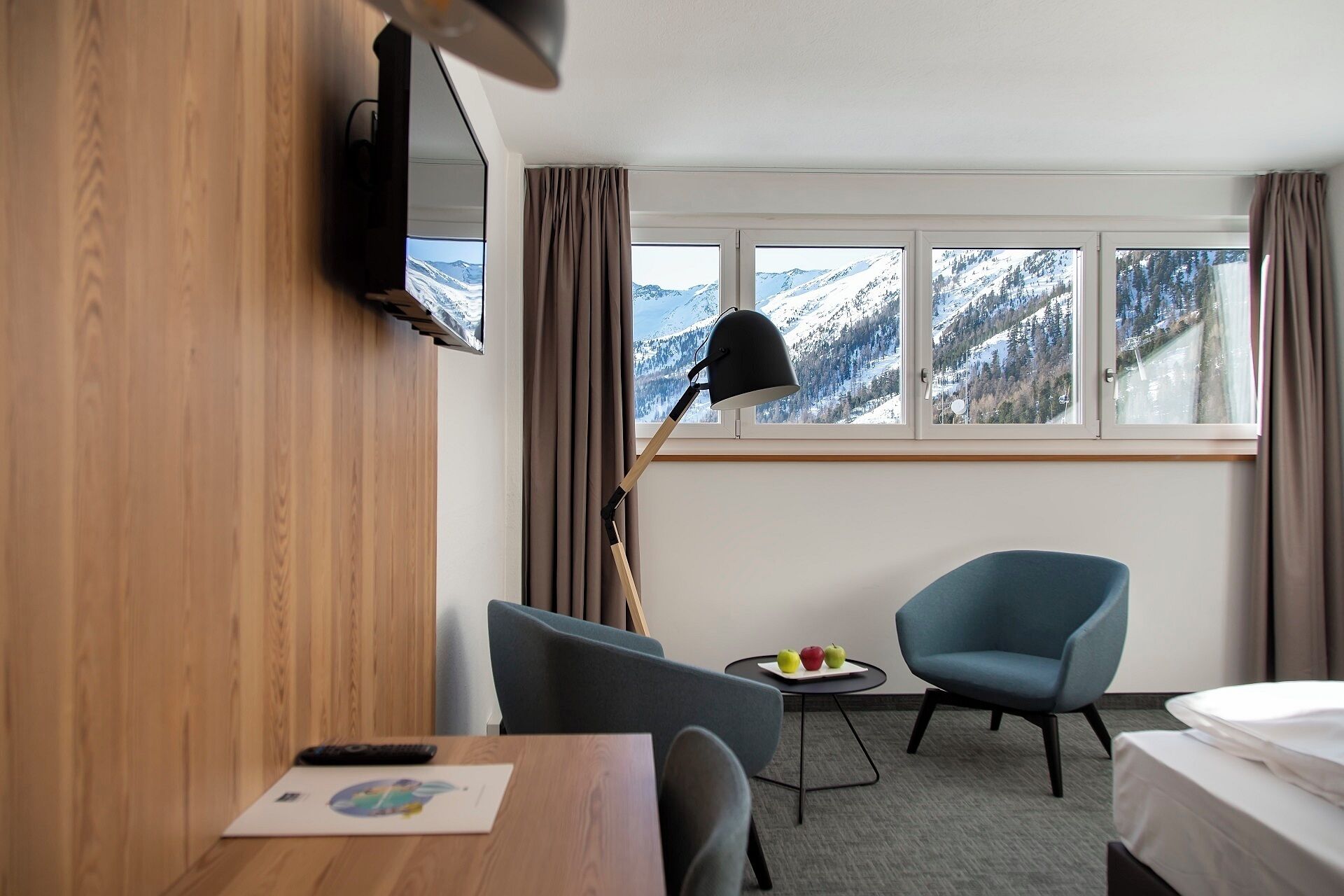 A hotel room with a bed, a desk with a remote control, a chair, a table with apples, a lamp, and a TV on the wall.
