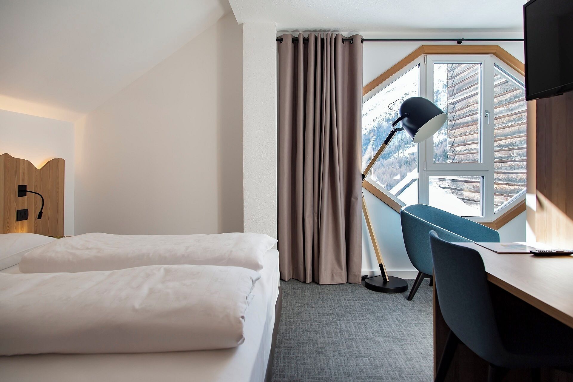 A hotel room with a bed, a desk, a chair, a lamp, and a window with curtains.