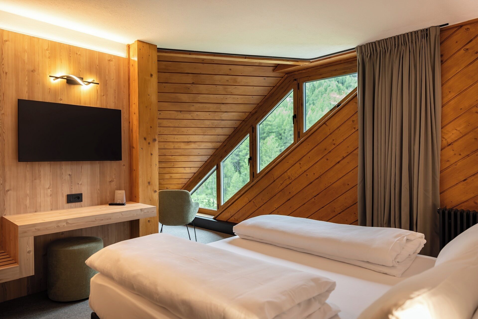 Une pièce dotée d’un plafond en pente en bois, d’un téléviseur à écran plat fixé au mur, d’un bureau avec une chaise et d’un lit recouvert de draps blancs impeccablement tendus.
