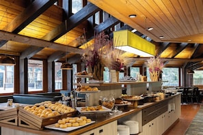 Een ontbijtbuffet met diverse gebakjes en brood, houten balken en grote ramen.