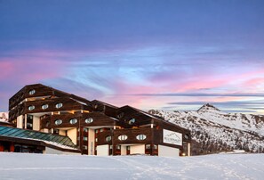 Een modern hotel met een uniek architectonisch ontwerp, meerdere verdiepingen en ronde ramen, gelegen voor een achtergrond van besneeuwde bergen en een kleurrijke lucht.