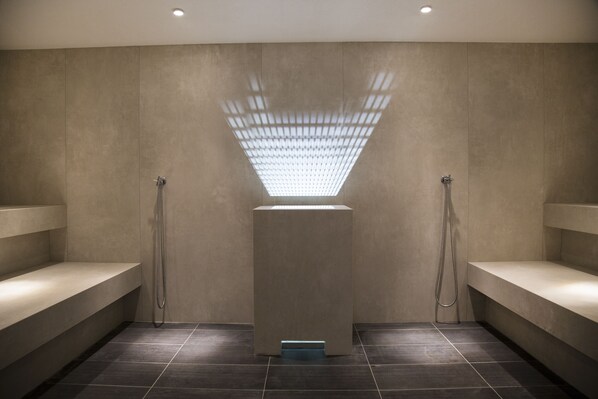 A modern shower area with a central light fixture, benches, and tiled flooring. - Blu Senales Active & Family Resort (Senales)