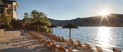 A beachside hotel with sun loungers and umbrellas, a paved walkway, and a view of the water and hills.