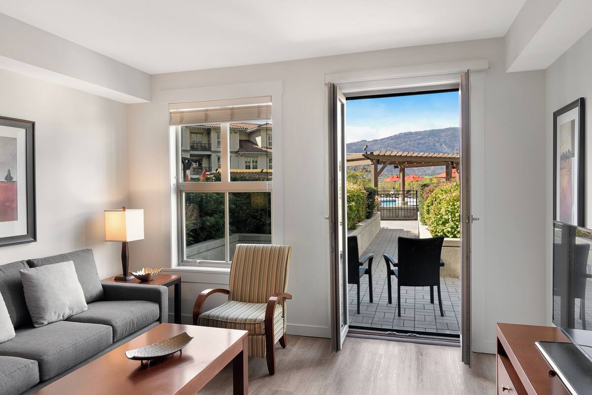 Un salon moderne avec un canapé gris, un fauteuil à rayures, une table basse en bois et un téléviseur posé sur un meuble en bois. Une grande fenêtre offre une vue sur une piscine et un espace de détente extérieur.