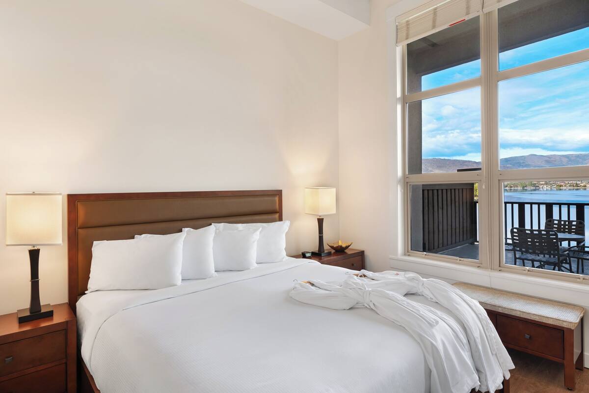 Une chambre d’hôtel dotée d’un grand lit, de deux tables de chevet avec des lampes, d’une vue sur l’extérieur grâce à une grande fenêtre et d’un balcon avec du mobilier d’extérieur.