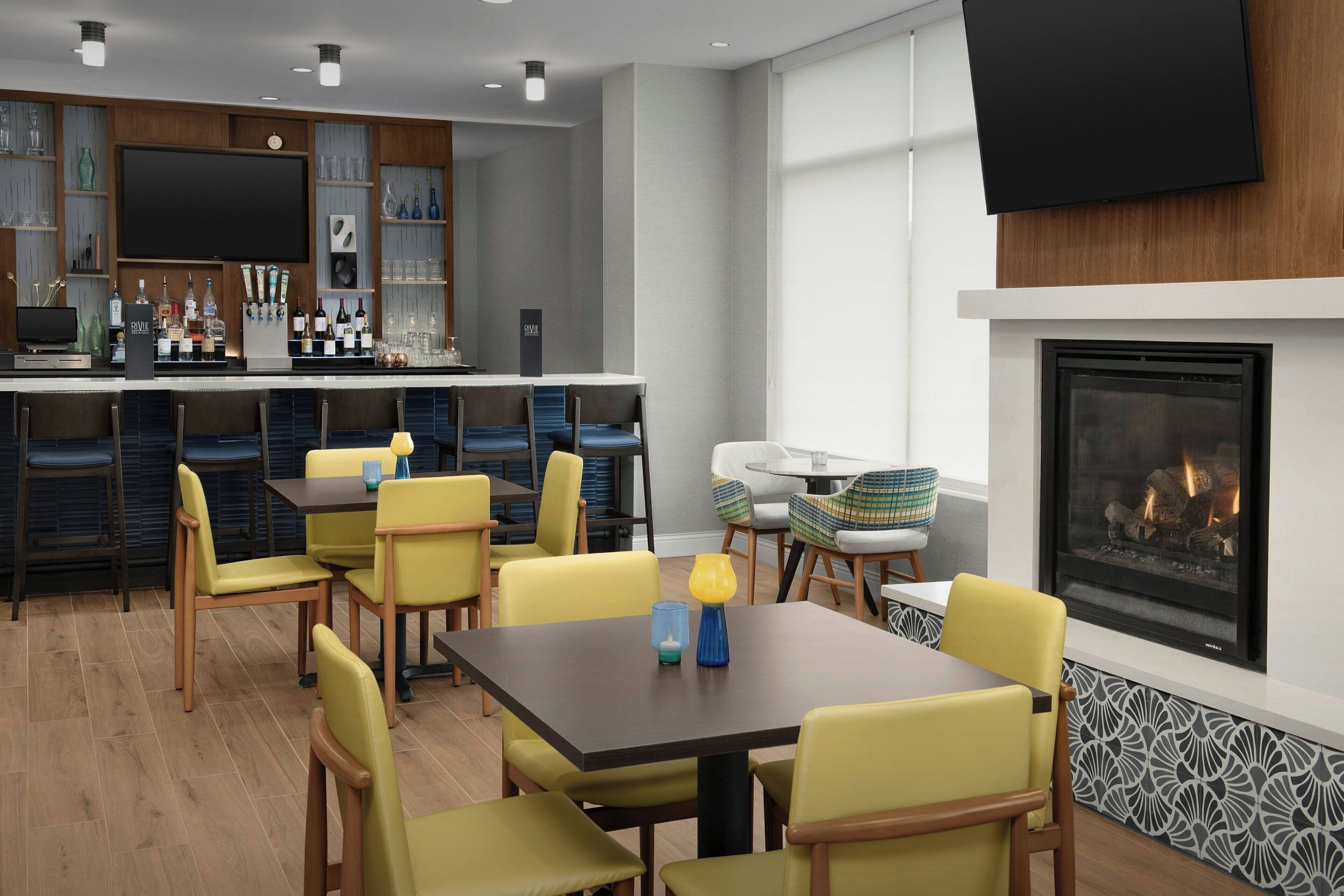 A modern bar area with a flat-screen TV, bar stools, and a variety of bottles. There are yellow chairs around a table with a blue candle and a small vase. A fireplace is visible in the background.