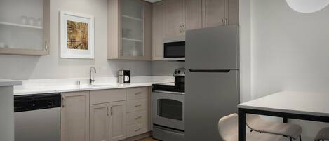 Una cocina moderna con armarios de madera clara, un refrigerador de acero inoxidable, un microondas y una cafetera. Hay una encimera blanca y una pequeña mesa de comedor con sillas.