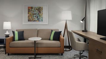 A hotel room with a sofa, a small round table, a desk with a chair, and a TV.