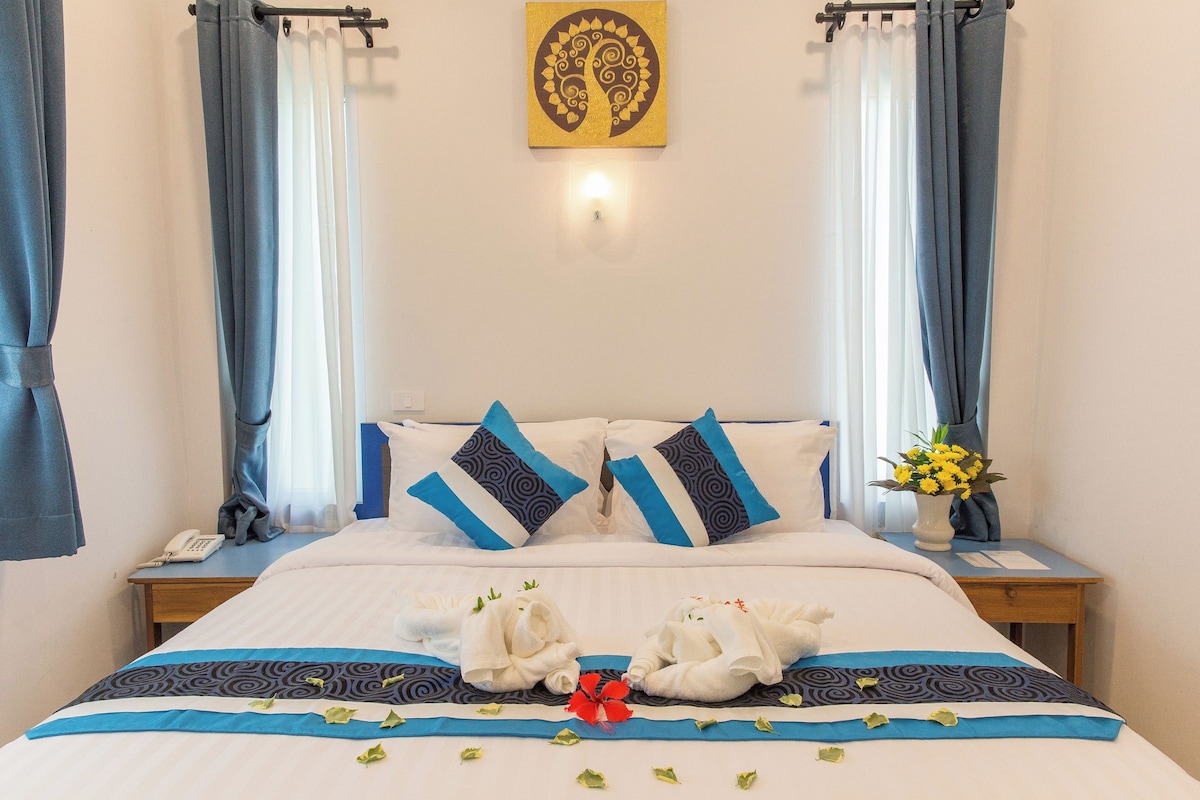 A hotel room with a bed, two pillows, a bedside table with a phone, a vase with flowers, and a wall-mounted artwork.