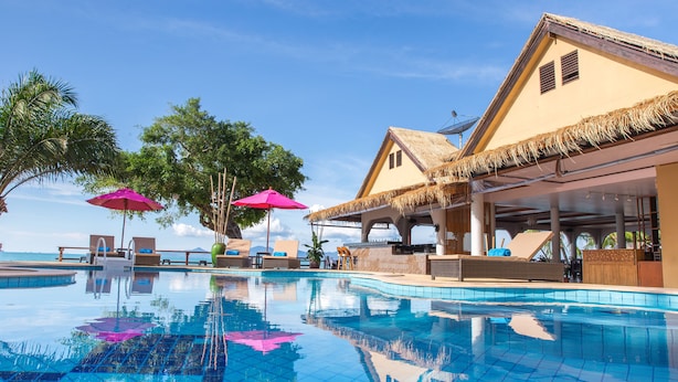A swimming pool with lounge chairs and umbrellas, surrounded by buildings with thatched roofs. - Adarin Beach Resort (Koh Samui)