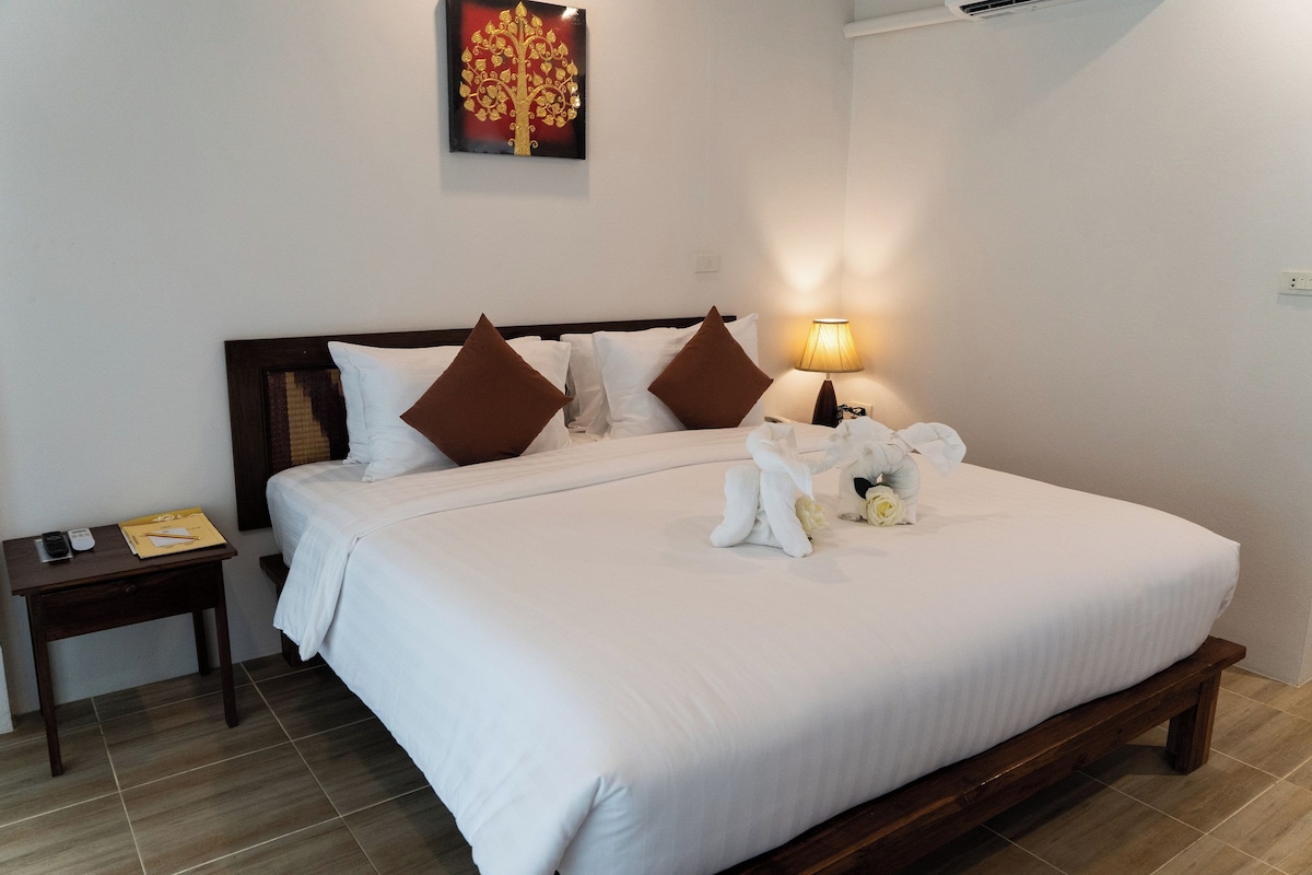 A neatly made bed with white linens and brown pillows, a bedside table with a lamp, and a wall-mounted artwork.