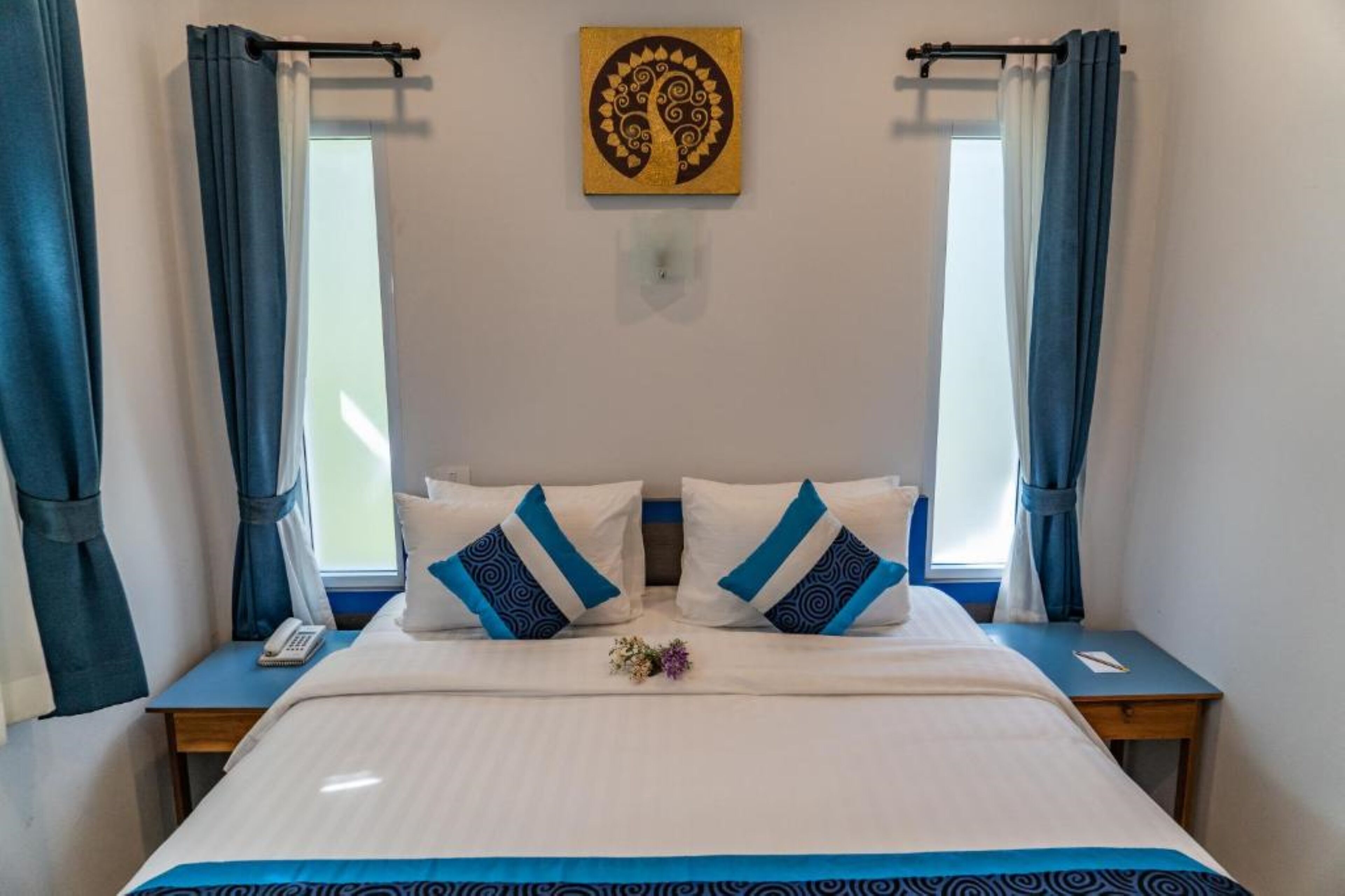 Una cama bien tendida con ropa de cama blanca y azul, dos mesitas de noche, una obra de arte en la pared y una ventana con cortinas.