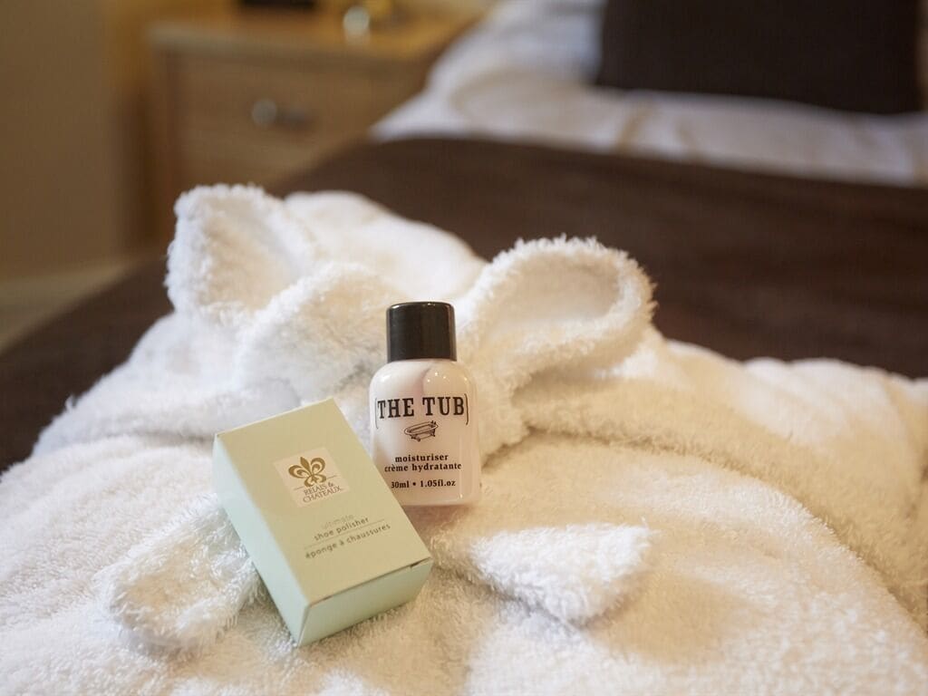 A hotel room with a bed covered in a white fluffy blanket, a bottle of 'The Tub' moisturizer, and a box of 'Bristol & Co.' soap.