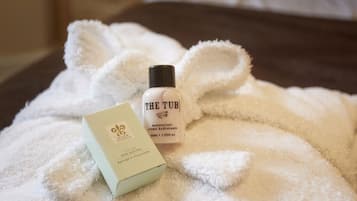 A hotel room with a bed covered in a white fluffy blanket, a bottle of 'The Tub' moisturizer, and a box of 'Bristol & Co.' soap.