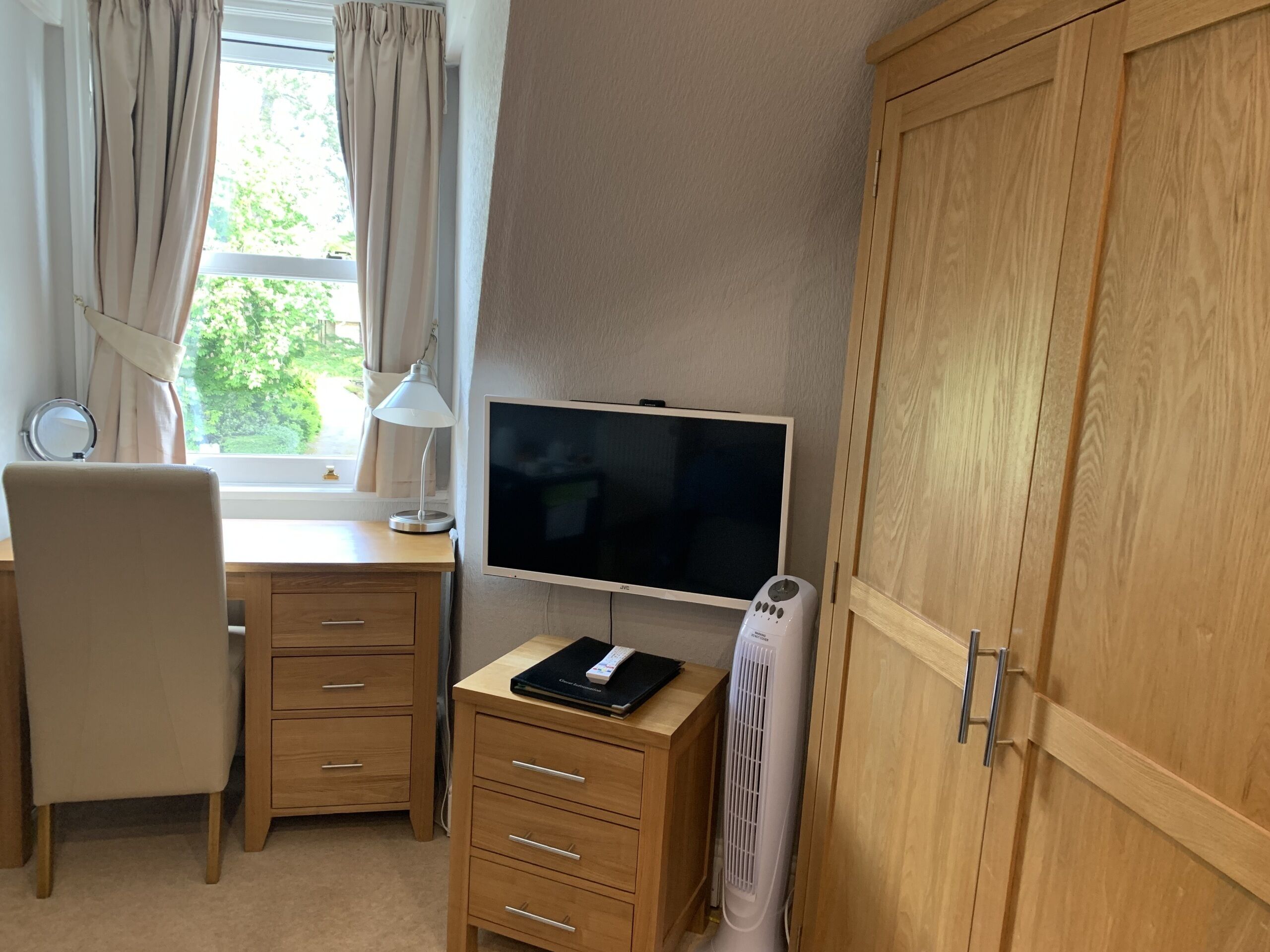 Een kamer met een houten bureau, een stoel, een televisie, een lamp, een verwarming en een kledingkast.
