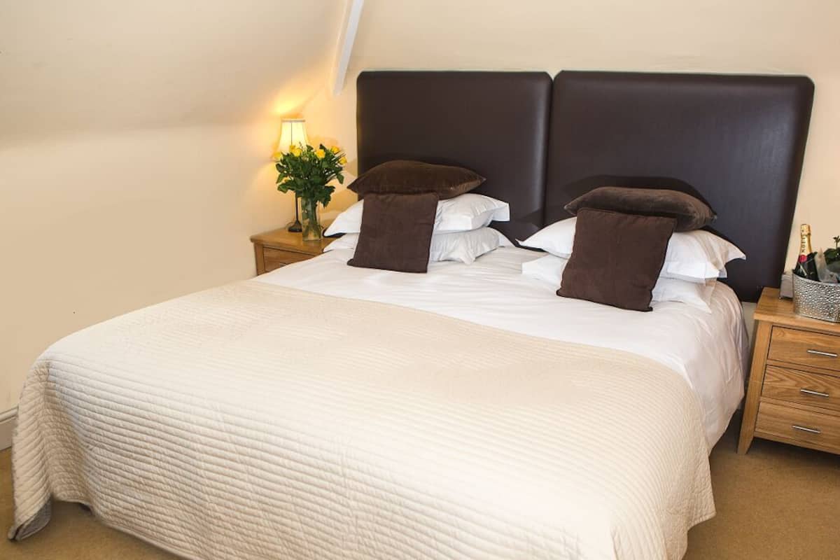 A neatly made bed with a white quilt, two dark brown pillows, and a dark headboard. There is a bedside table with a lamp and a vase of flowers.
