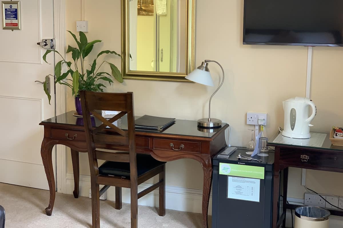 A hotel room with a wooden desk, a chair, a potted plant, a desk lamp, a kettle, a trash can, a mirror, and a television.