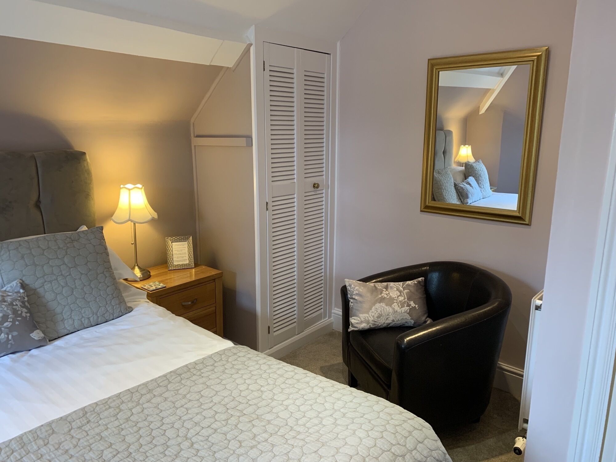 Een hotelkamer met een bed, een stoel, een nachtkastje met lamp, een kledingkast en een spiegel aan de muur.