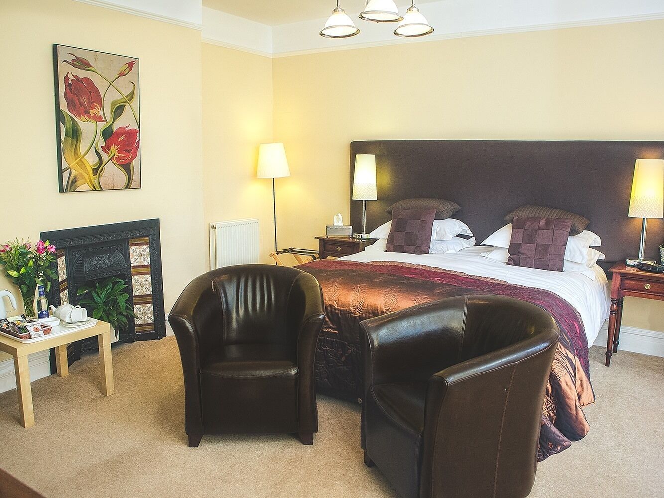 Una habitación de hotel con una cama grande, dos mesitas de noche con lámparas, una chimenea, una pequeña mesa con un jarrón de flores y dos sillones de cuero.