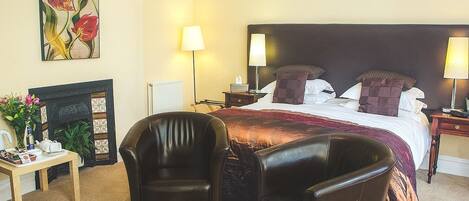 Una habitación de hotel con una cama grande, dos mesitas de noche con lámparas, una chimenea, una pequeña mesa con un jarrón de flores y dos sillones de cuero.